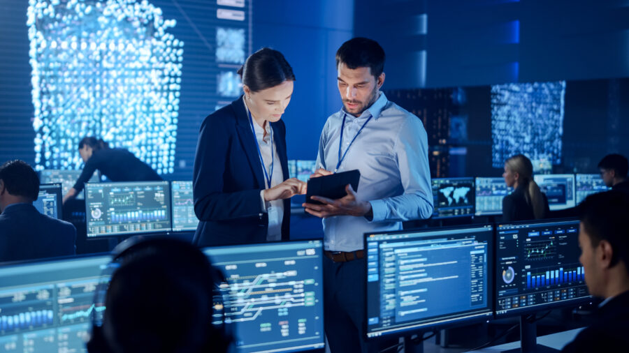 Man and women looking at a tablet, in a room full of people working on computers.
